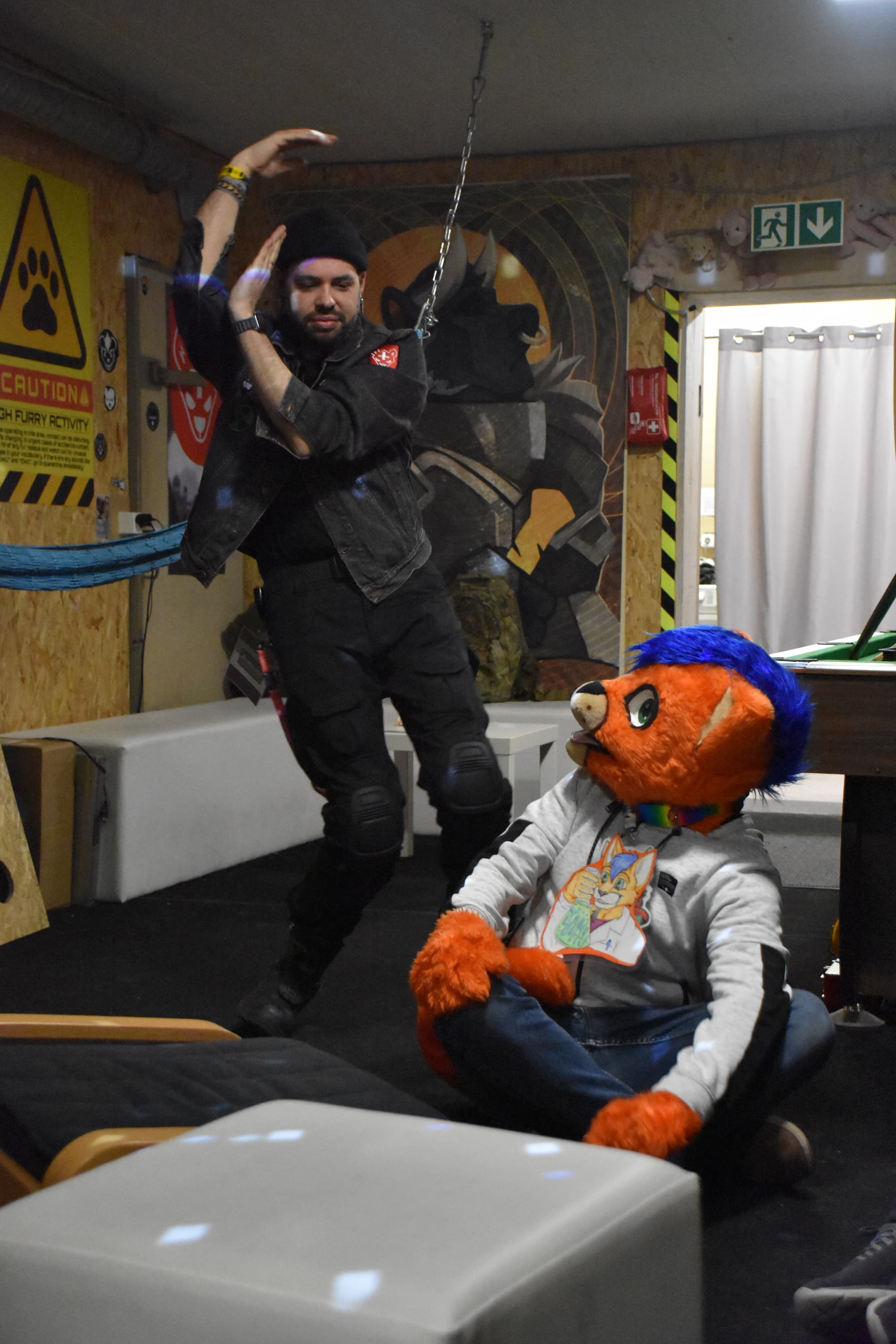 A man in black clothing dancing, caught doing a pose somewhat looking like an "S", a person in an orange cat fursuit is looking amazed at the performed dance.
